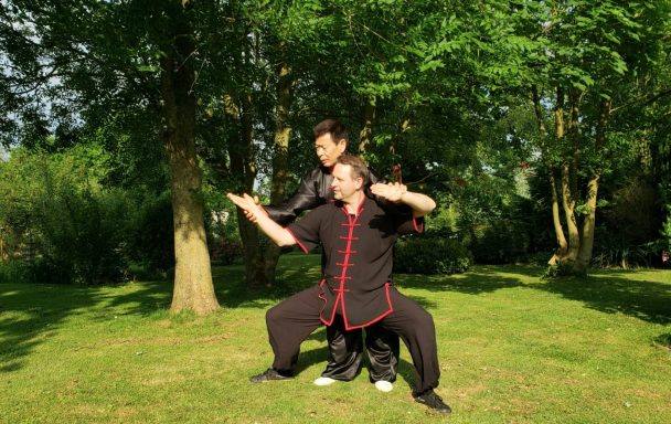 Zwei Personen in schwarzer Kleidung führen eine Tai-Chi-Übung im Freien aus.