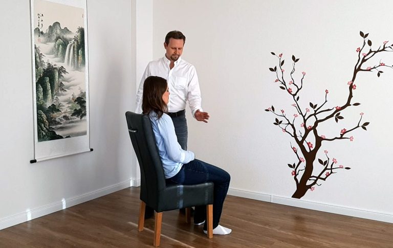 Ein Mann spricht mit einer sitzenden Frau vor einer Wand mit einem Baum-Motiv.