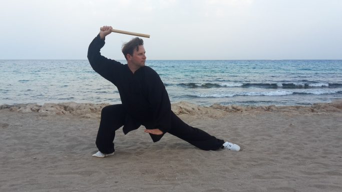 Kämpfer in schwarzer Kleidung übt mit einem Stock am Strand.