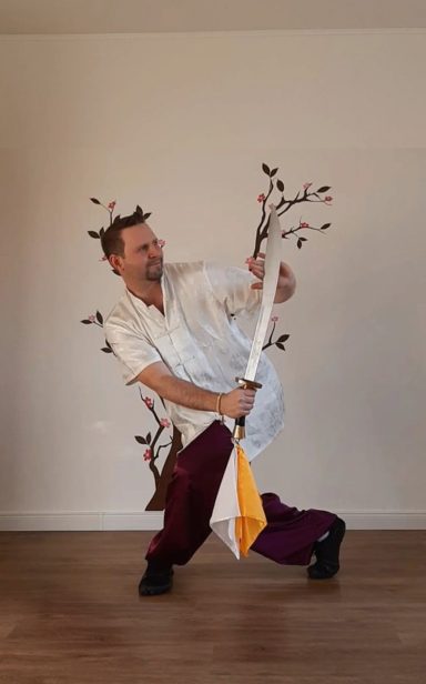 Mann in weißem Oberteil und lila Hose, hält einen langen Stock, umgeben von Ästen.