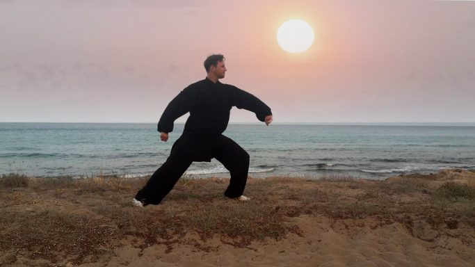 Person in schwarzer Kleidung übt Tai Chi am Strand bei Sonnenuntergang.