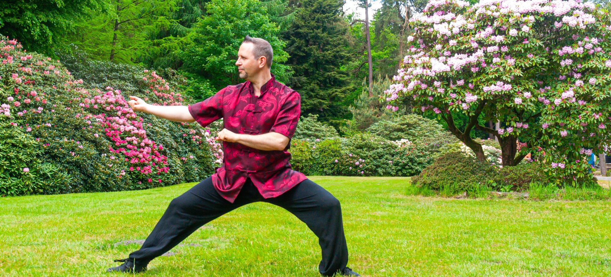 Mann in rot-schwarzem Kimono übt Tai Chi im Garten mit blühenden Bäumen.