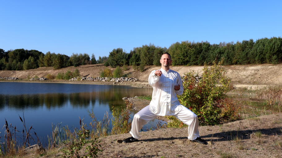 Eine Person in weißer Kleidung übt Tai Chi am Ufer eines ruhigen Gewässers.