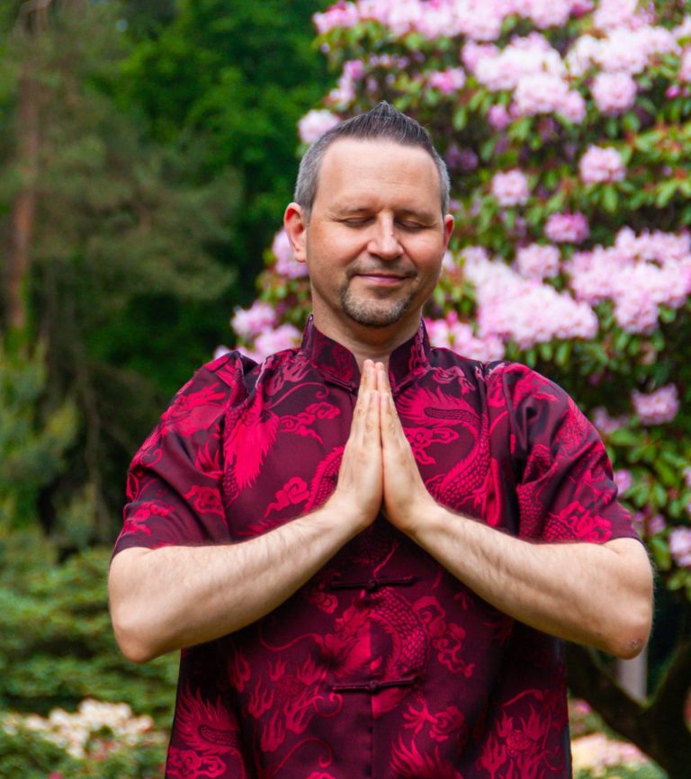 Meditation Mann im roten traditionellen Outfit mit gefalteten Händen vor blühenden Pflanzen.