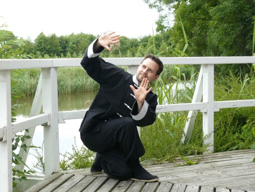 Mann in schwarzer Martial-Arts-Kleidung vor einem Wasserlauf in einer natürlichen Umgebung.