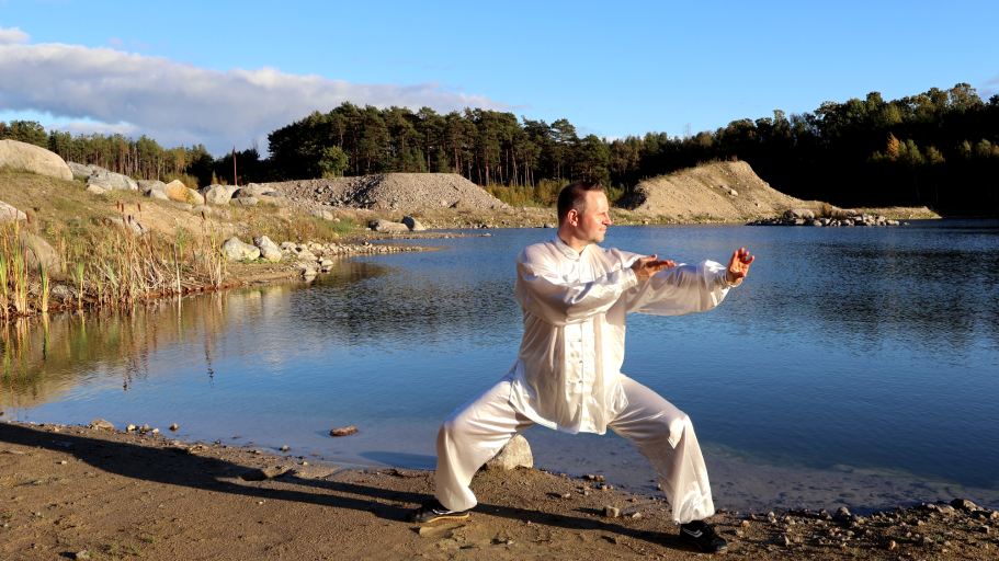 Mann in traditioneller Kleidung übt Tai Chi an einem Seeufer bei Sonnenuntergang.