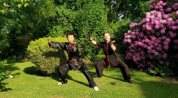 Zwei Personen in traditioneller Kleidung üben Kung Fu in einem grünen Garten.