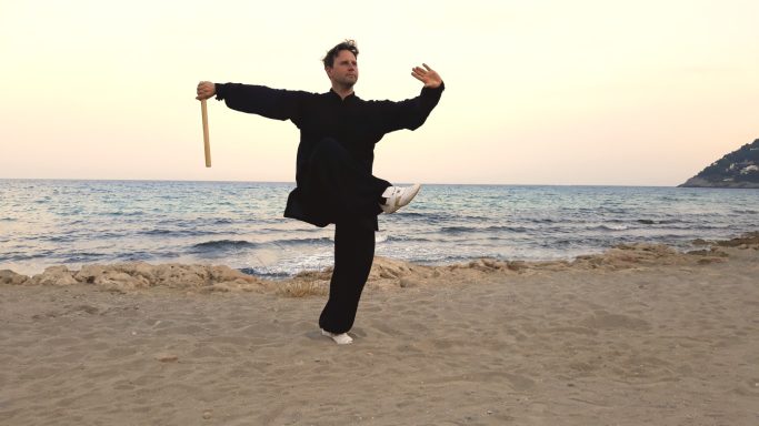 Ein Mann in schwarzer Kleidung übt mit einem Stock am Strand bei Sonnenuntergang.