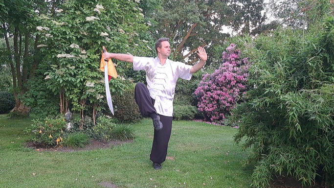 Mann in martial-arts-Kleidung führt eine Bewegung mit einem Schwert im Garten aus.