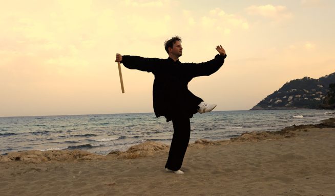 Person in schwarzem Outfit übt Kampfkunst am Strand bei Sonnenuntergang.