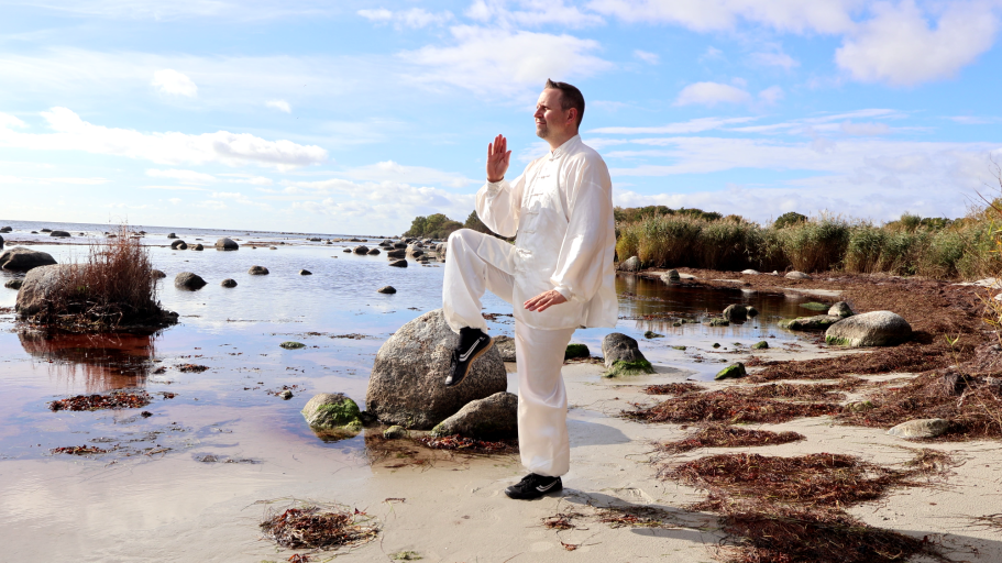Mann in weißer Kleidung übt Tai Chi am Strand mit Felsen und Wasser im Hintergrund.